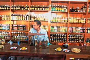 In einem Geschäft mit holzgetäfelten Regalen, die bis zur Decke mit Flaschen gefüllt sind, steht ein Mann hinter einer langen Verkaufstheke. Er ist gerade dabei, vier verschiedene Mezcal-Flaschen für eine Verkostung vorzubereiten. Auf der Theke stehen kleine Teller mit Orangenscheiben und anderen Beilagen. Die Szene vermittelt den Eindruck einer traditionellen Mezcal-Verkostung in Mexiko.