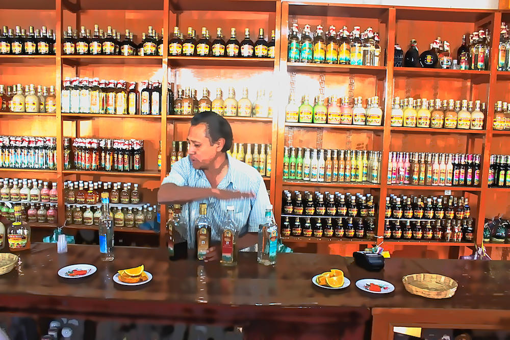 In einem Geschäft mit holzgetäfelten Regalen, die bis zur Decke mit Flaschen gefüllt sind, steht ein Mann hinter einer langen Verkaufstheke. Er ist gerade dabei, vier verschiedene Mezcal-Flaschen für eine Verkostung vorzubereiten. Auf der Theke stehen kleine Teller mit Orangenscheiben und anderen Beilagen. Die Szene vermittelt den Eindruck einer traditionellen Mezcal-Verkostung in Mexiko.