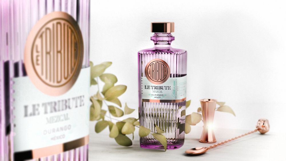 Elegantes Produktfoto einer geriffelten, leicht violett schimmernden Mezcal-Flasche „LE Tribute Mezcal Durango, Mexico“ mit goldfarbenem Verschluss und rundem Logo auf dem Etikett. Daneben liegen Barzubehör aus Roségold (Jigger und Barlöffel) auf einer hellen Oberfläche, im Hintergrund dekorative grüne Zweige und links unscharf eine passende Verpackungsbox derselben Marke. Helles, weiches Licht sorgt für eine hochwertige, stilvolle Atmosphäre.