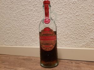 Eine Nahaufnahme der Flasche „El Ron Prohibido“, eines mexikanischen Rums. Die Flasche ist aus klarem Glas gefertigt und hat eine rustikale Form mit einem kleinen Henkel am Hals. Sie trägt ein auffälliges, rotes und bronzefarbenes Etikett, das den Namen und Angaben wie „Solera 12“ sowie „Mexican Rum“ enthält. Der Hals ist mit einem roten Siegel und einer roten Kapsel verschlossen. Die Flasche steht auf einem hellen Holzboden vor einer weißen, strukturierten Wand.