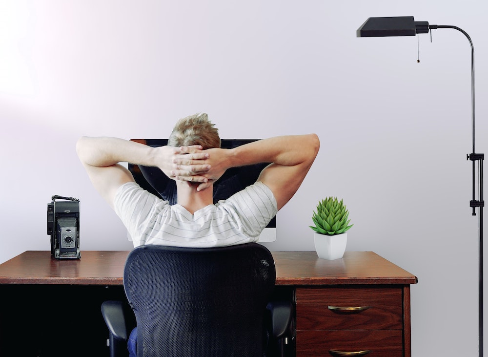 „Ein Mann mit kurzen blonden Haaren sitzt mit dem Rücken zum Betrachter in einem schwarzen Bürostuhl. Er trägt ein weiß gestreiftes T-Shirt und hat seine Hände entspannt im Nacken verschränkt. Er blickt auf einen Computerbildschirm auf einem dunklen Holztisch. Auf dem Schreibtisch stehen links eine alte Vintage-Kamera und rechts eine kleine grüne Pflanze in einem weißen Topf. Rechts im Bild ragt eine schwarze Stehlampe empor. Der Hintergrund ist eine schlichte, helle Wand.“