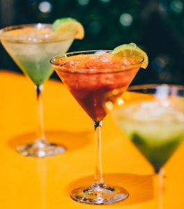„Drei farbenfrohe Cocktails in Martinigläsern mit Limettengarnitur auf einer gelben Bartheke.“