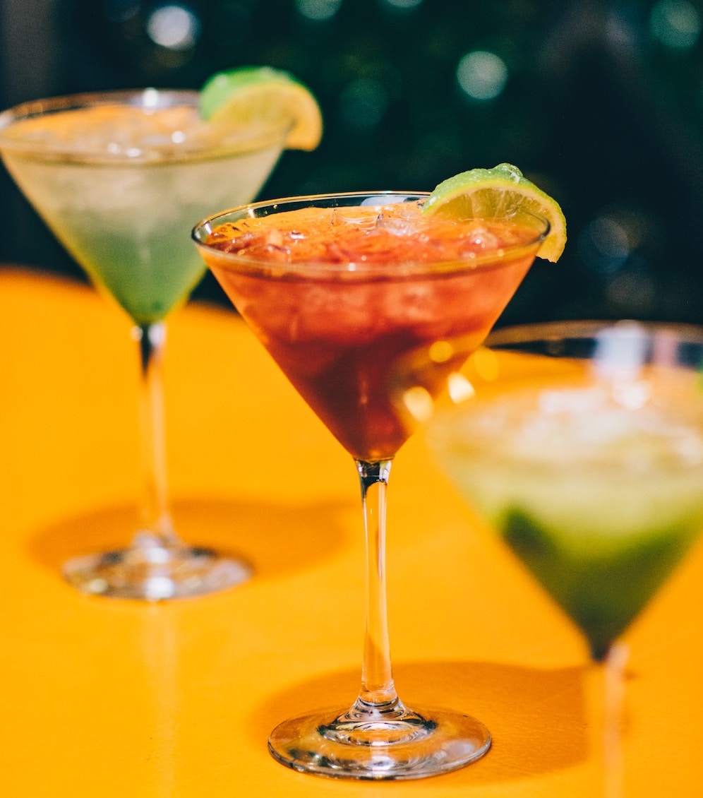 „Drei farbenfrohe Cocktails in Martinigläsern mit Limettengarnitur auf einer gelben Bartheke.“