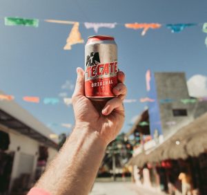 „Eine Hand hält eine Dose Tecate Original Bier vor blauem Himmel mit bunten Wimpelketten.“