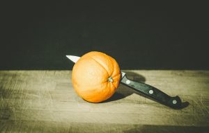 „Eine ganze Orange, die von einem schwarzen Küchenmesser durchstochen auf einem Holzbrett liegt.“