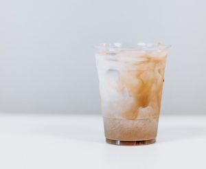 „Ein durchsichtiger Kunststoffbecher mit einem Kaltgetränk vor einem neutralen, hellgrauen Hintergrund. Im Becher vermischt sich weißliche Milch in Schlieren mit hellbraunem Kaffee. Man erkennt Eiswürfel im oberen Bereich des Getränks. Das Bild ist minimalistisch und hell gestaltet.“