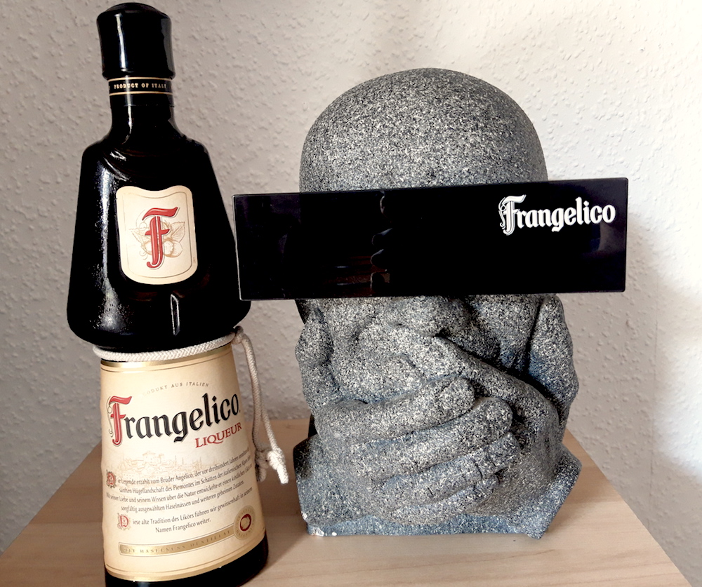 „Produktfoto auf einem hellen Holztisch: Links steht eine markante dunkle Glasflasche des italienischen Likörs Frangelico mit hellem Etikett und weißer Kordel. Rechts daneben steht eine graue Büste aus steinartigem Material, deren Hände den Mund bedecken. Über den Augen der Skulptur liegt ein breiter, schwarzer Balken (wie eine Sonnenbrille), auf dem in weißer Schreibschrift der Markenname 'Frangelico' steht.“