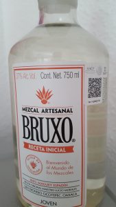 Eine Frontalansicht einer klaren Glasflasche mit dem weißen Etikett des „Bruxo“ Mezcal Artesanal. Das Etikett ist rot umrandet und zeigt oben die Angaben „37% Alc. Vol.“ und „Cont. Net. 750 ml“. Zentral im Fokus steht das schwarze Markenlogo „BRUXO“ unter einem roten Agaven-Symbol. Im unteren Bereich finden sich weitere Details wie „100% MAGUEY ESPADÍN“, der Name des Maestro Mezcalero sowie der Herkunftsort „San Dionisio Ocotepec, Oaxaca“ und die Sortenbezeichnung „JOVEN“.